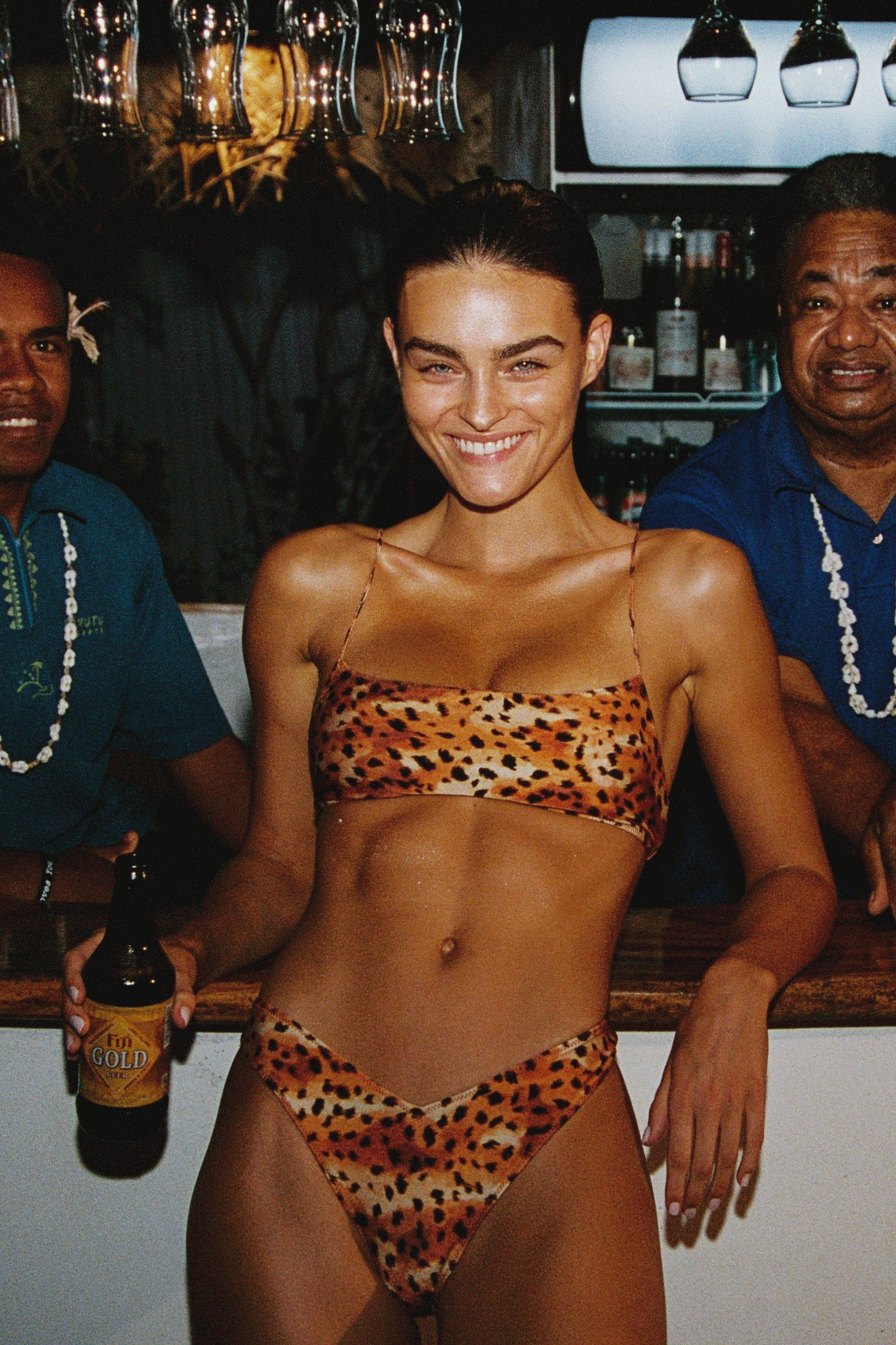 Woman in leopard print bikini holding a beer at a bar with two men behind her.