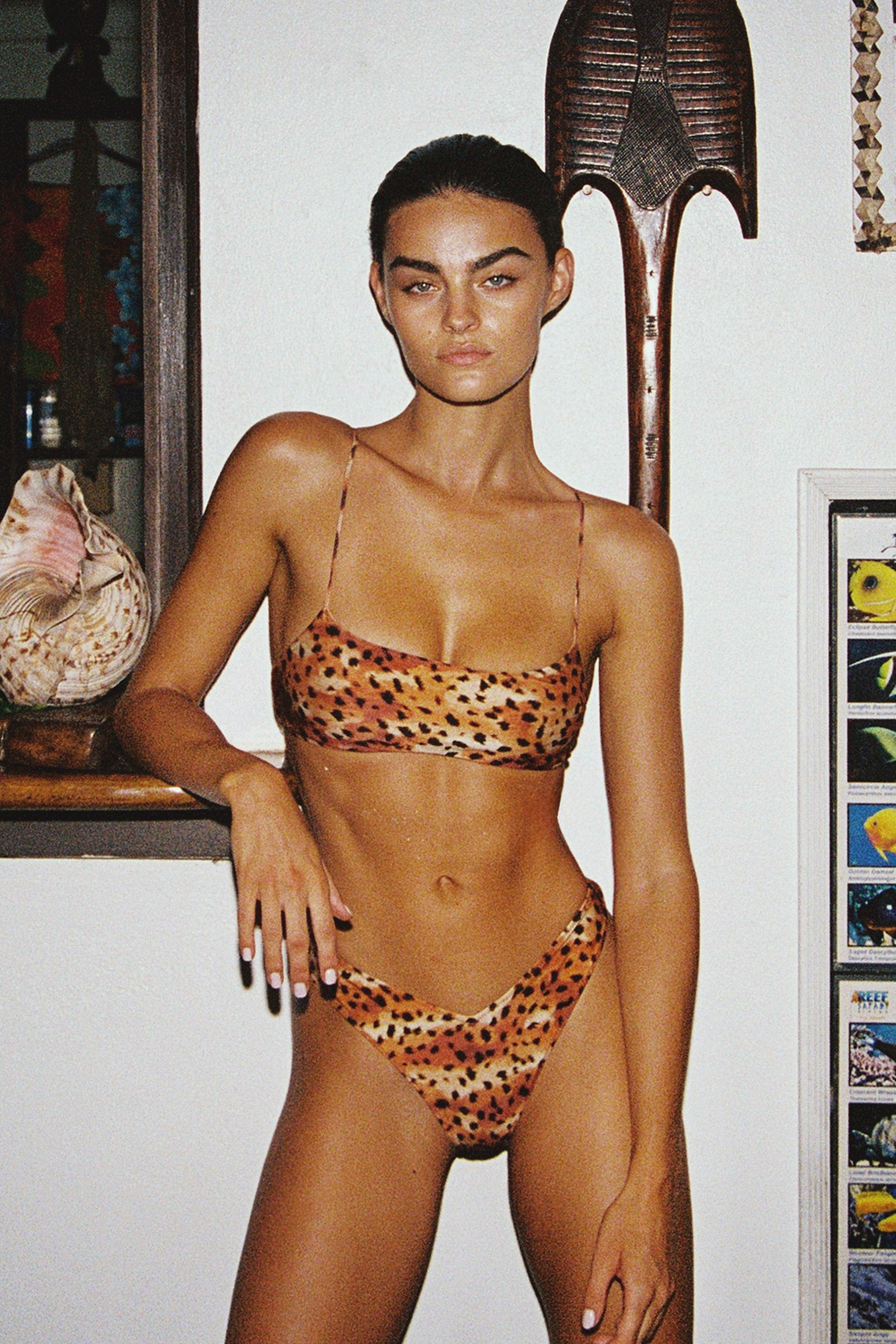 Woman wearing a leopard print bikini in an indoor setting with a shelf and decorative items.