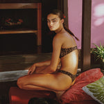 Woman in leopard print bikini sitting on a red couch with a dog, against a pink wall.