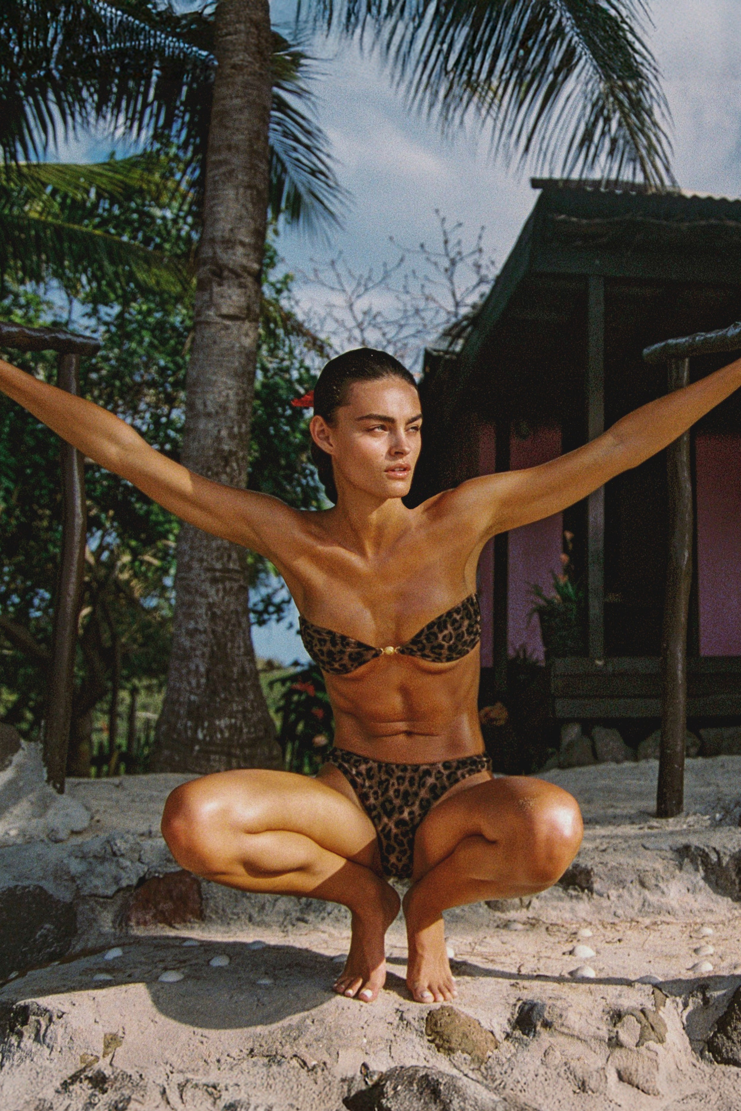 Person in a leopard print bikini posing outdoors with palm trees and a hut in the background